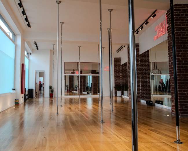 The image shows a spacious studio with wooden flooring, pole dance poles arranged evenly, and large windows allowing natural light to enter.