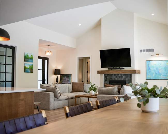 The image shows a modern living room with a large sectional sofa, a mounted television above a fireplace, and a dining area featuring a wooden table with chairs.