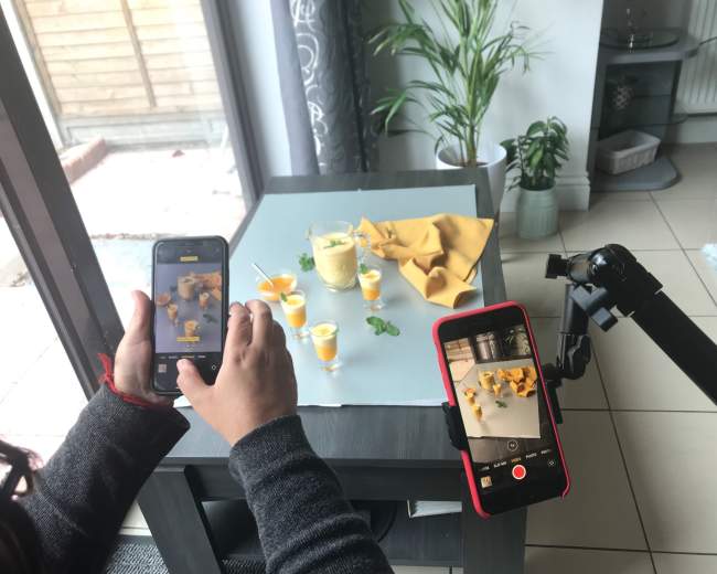 A person is taking photos and videos of small glasses filled with a yellow drink on a table, with a plant in the background.