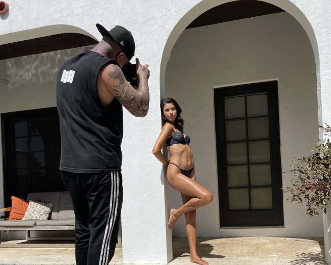 A photographer captures a model in a bikini posing on steps beside a pool at a modern house.