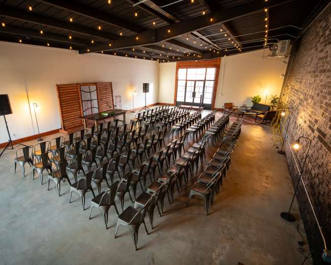 A spacious event venue features rows of metal chairs arranged for a gathering, with string lights illuminating the ceiling.