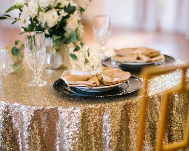 A table is set with elegant plates and napkins, surrounded by decorative glassware and a floral centerpiece, all draped with a sparkling gold tablecloth.