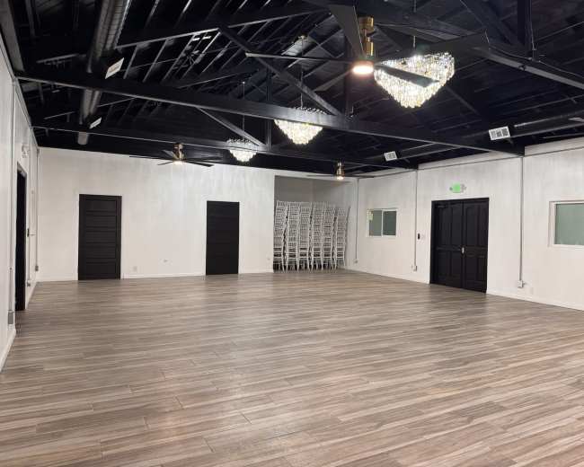 The image shows a spacious, well-lit room with a sleek wooden floor, black ceiling beams, and two ornate chandeliers.