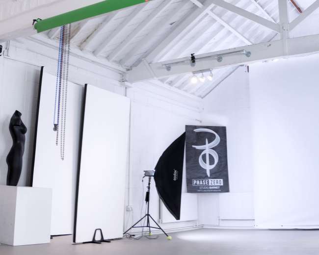 The image shows a spacious photography studio with a large clock on the wall, a green beam overhead, and various photography equipment including a softbox and backdrop.