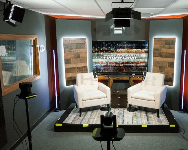 The image shows a television studio setup with two chairs, a small table, and various lighting equipment arranged around a wooden backdrop.