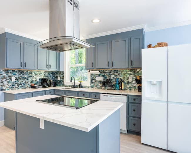 A modern kitchen features grey cabinetry, a large island with a marble countertop, and a stainless steel range hood.