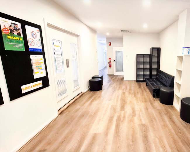 The image shows a modern, well-lit corridor with seating areas, shelves, and a large television mounted on the wall.