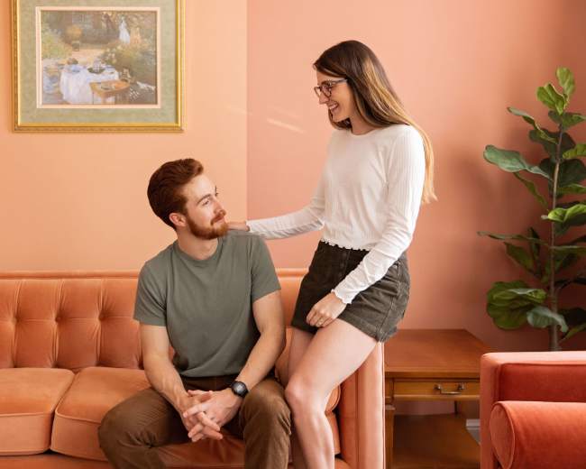 A man sits on a peach-colored couch while a woman stands beside him, both smiling in a softly lit, pastel-hued room.