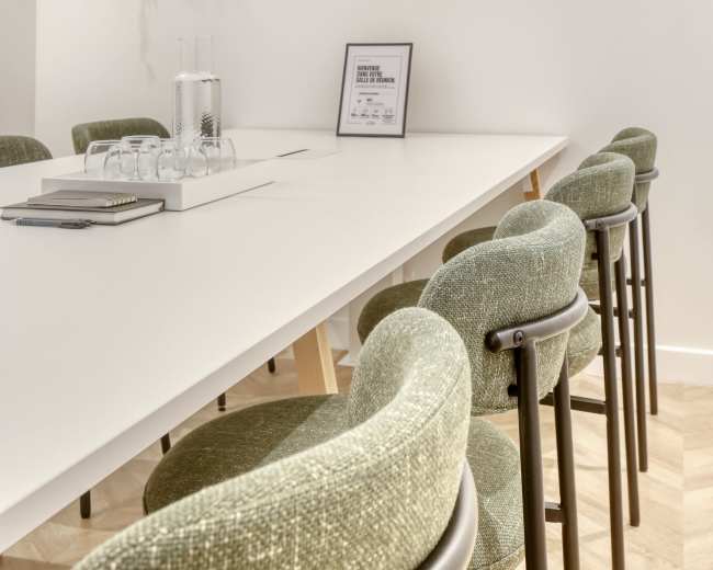 A long white table with green upholstered chairs is set in a modern meeting room decorated with wooden shelves and plants on the walls.