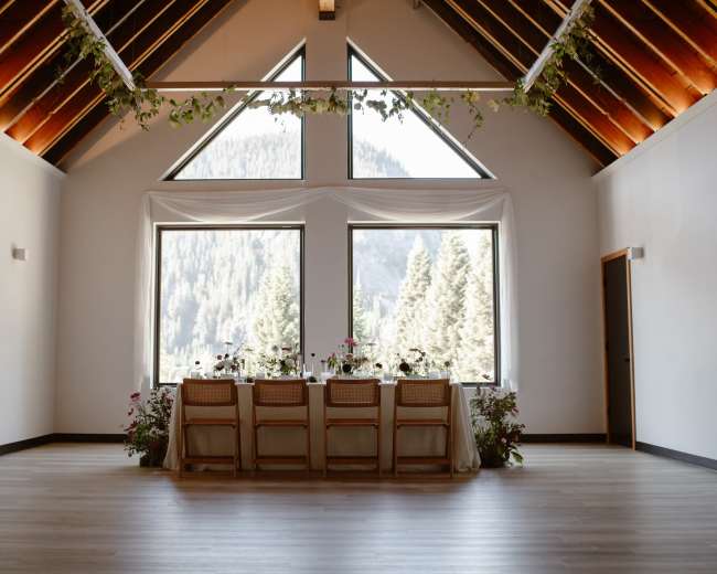 A long dining table with floral arrangements is set against large windows that showcase a forested mountain landscape.