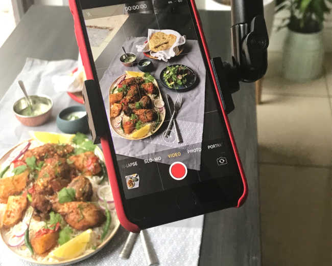 A smartphone is mounted on a tripod, recording a table set with a platter of food, including grilled meat and a side salad.