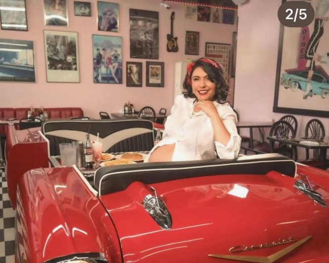 A woman seated in a vintage red convertible inside a 1950s diner, with retro decor and checkered floors in the background.