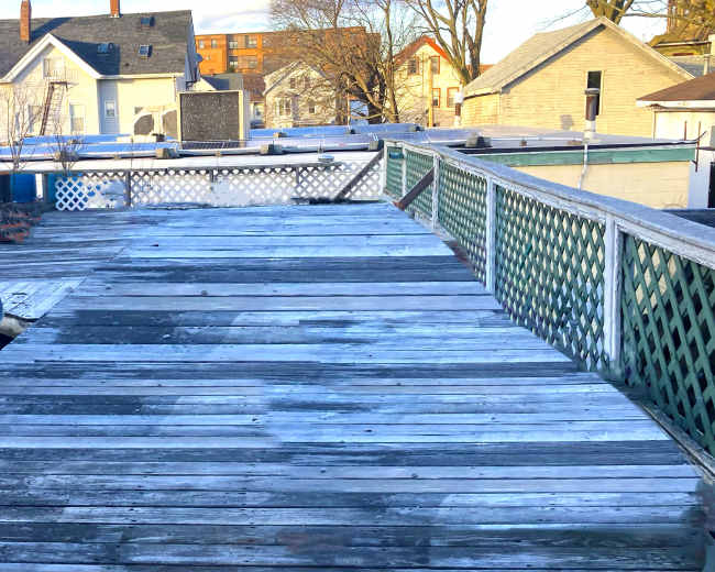 A weathered wooden deck leading toward a series of houses under a blue sky with scattered clouds.