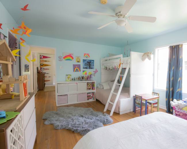 A light blue children's bedroom with a loft bed, a small table and chairs, and colorful decorations on the walls.