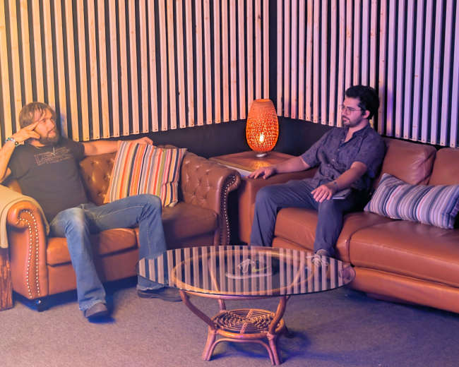 Two men are seated on a pair of leather sofas in a warmly lit room, with decorative lighting and a glass coffee table between them.