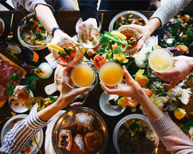 A group of hands raises various drinks in a toast above a table set with food and floral decorations.