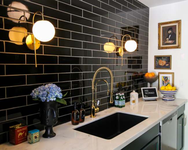 The image shows a modern kitchen with black tiled walls, a white countertop, a sink with a gold faucet, and decorative items including fruit and artwork.