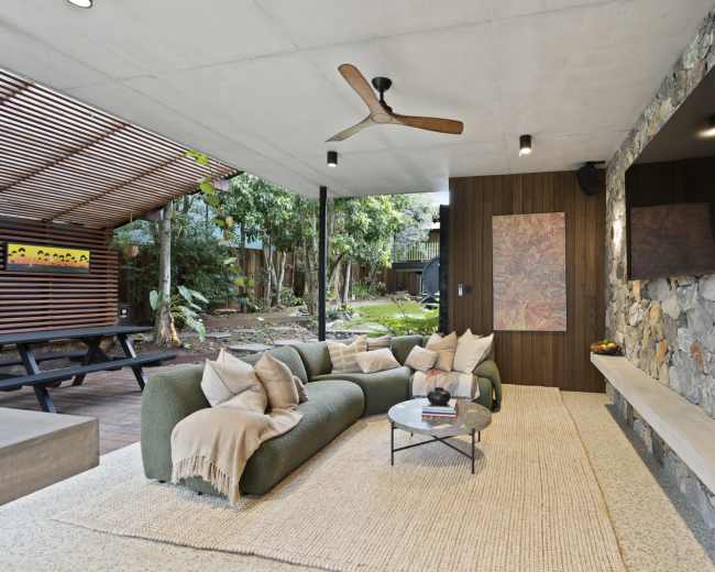 The image depicts a modern outdoor living space featuring a stone accent wall, comfortable seating, and a coffee table, surrounded by lush greenery.