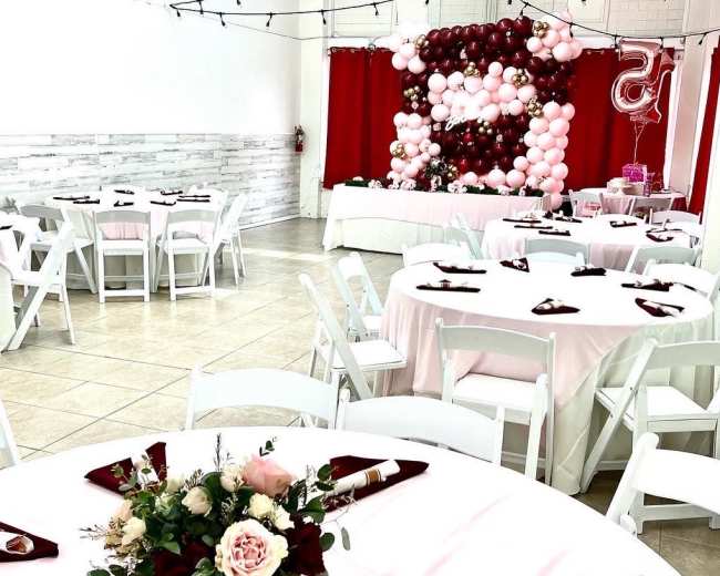 A decorated event space features round tables with white linens and maroon napkins, surrounded by a backdrop of balloons and a large "5" display.