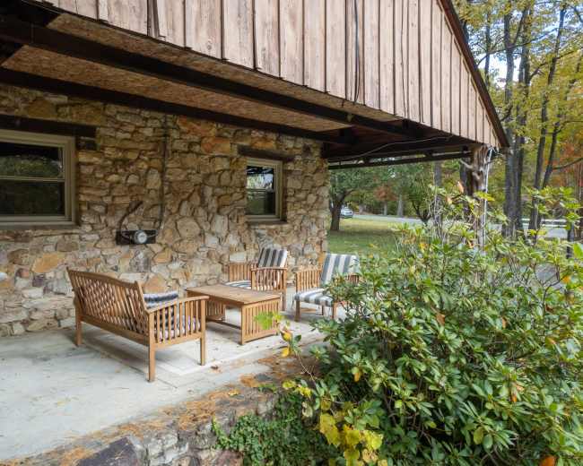 A rustic stone patio features wooden seating and a coffee table beneath an overhanging roof.