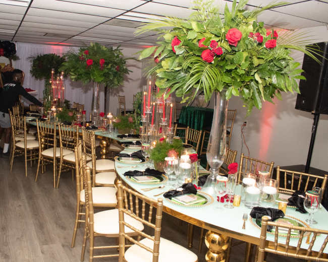 A long, elegantly set banquet table adorned with greenery and red roses is arranged in a decorated room featuring black balloons and a DJ setup.