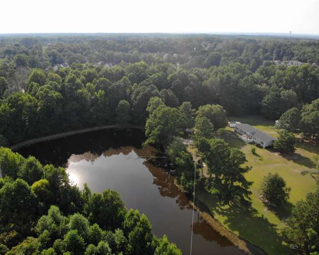 The image shows an aerial view of a circular pond surrounded by dense greenery and a single house located near the water's edge.