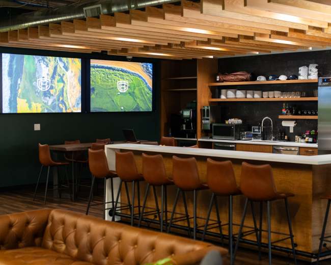 The image shows a modern kitchen and lounge area with bar seating, a stainless steel refrigerator, and two large screens displaying images on the wall.