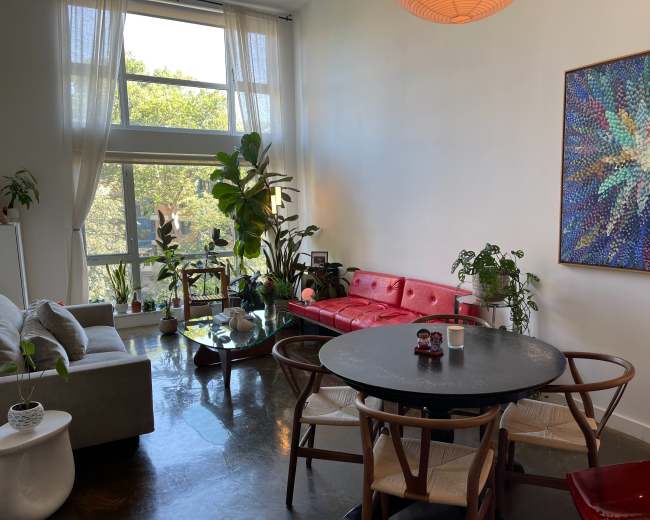 The image shows a modern living space with a red couch, a round dining table, and various indoor plants by large windows that provide natural light.