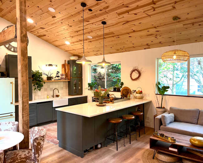 The image shows a modern kitchen and living area featuring wooden ceilings, a large central island with bar stools, green cabinetry, and ample natural light from a large window.