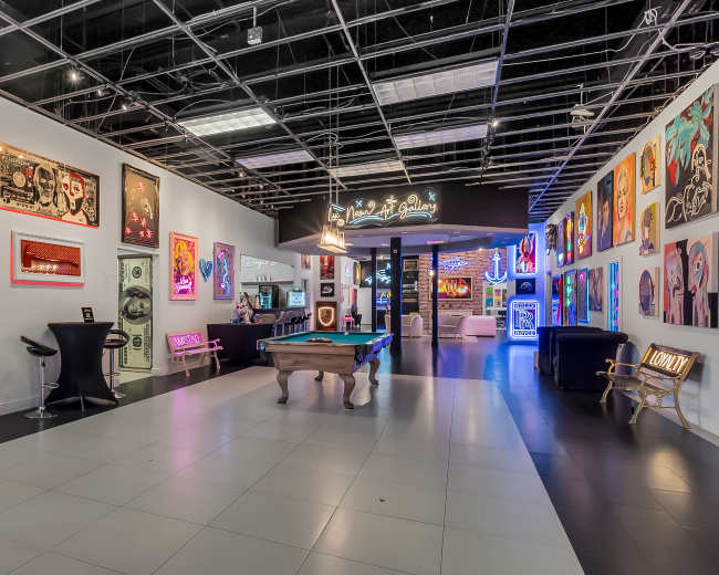 The image features a spacious art gallery with vibrant wall art, a pool table in the center, and an illuminated entrance leading to another area.