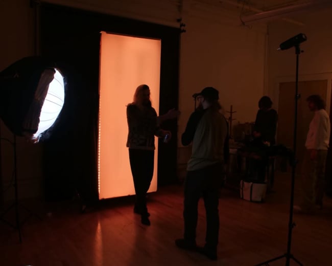 A person with long hair interacts with another individual in a photography studio illuminated by an orange backdrop and lighting equipment.