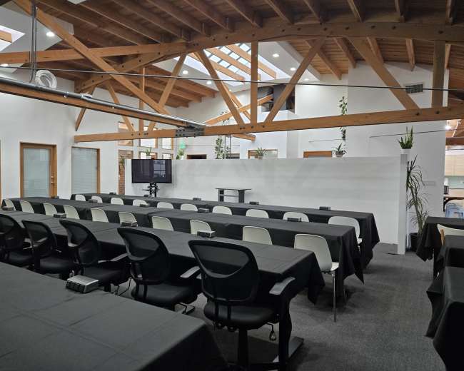 The image shows a modern conference room with multiple black tables and chairs arranged in rows, featuring a large screen and wooden beams overhead.
