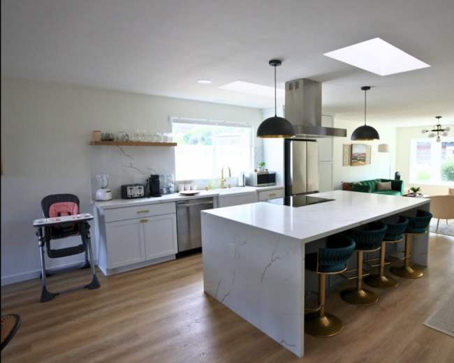 The image depicts a modern kitchen with a large white island, green high-backed bar stools, and a well-lit dining area featuring a sofa and armchairs.