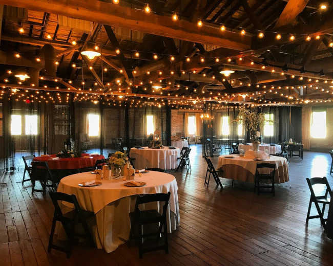 A spacious event hall with round tables set for dining, adorned with string lights overhead and large windows allowing natural light to enter.
