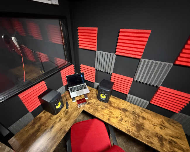 The image shows a small studio equipped with two speakers, a laptop, and soundproofing panels in red and gray on the walls.