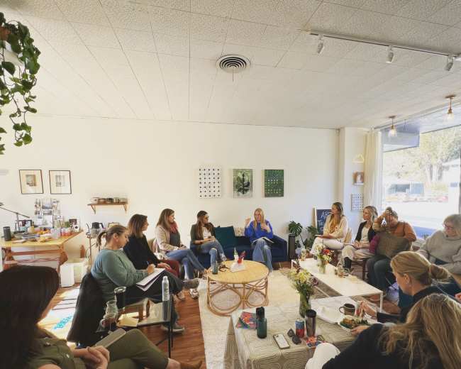 A group of women sits in a bright, open space, engaging in conversation while surrounded by plants and minimalist decor.