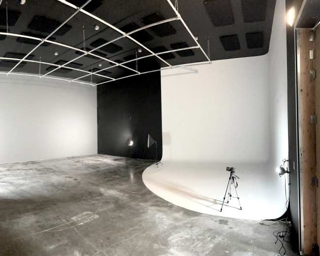A spacious photography studio with a black wall on one side and a white backdrop on the other, along with lighting equipment and tripods set up.