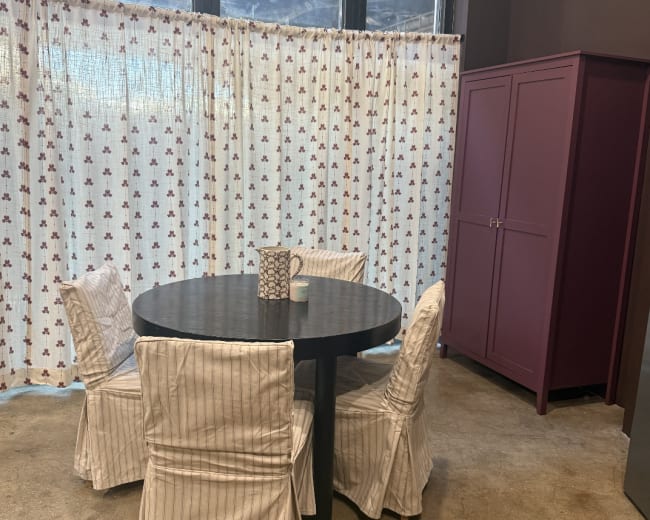 A round black table is set with four white striped chairs in a room with a curtain and a large purple cabinet.