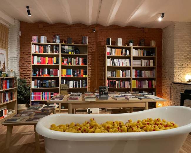 The image shows a cozy bookshop featuring bookshelves filled with various books, a central table with more books, and a bathtub filled with rubber ducks.