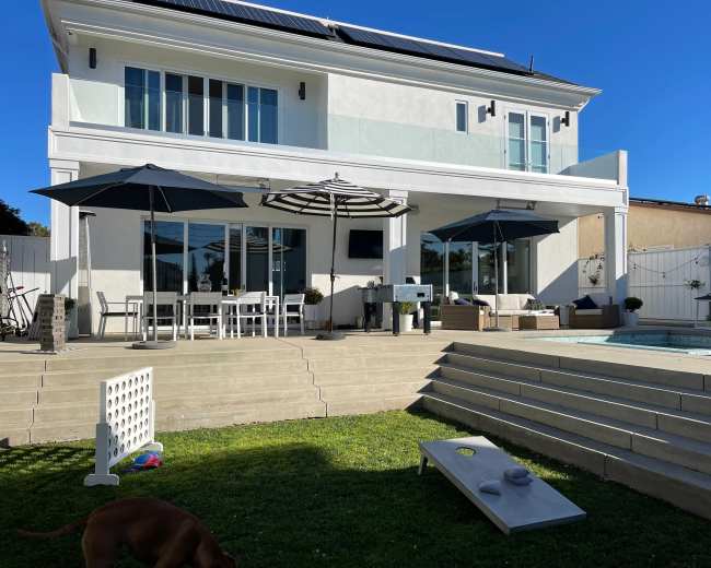 The image shows a modern two-story house with a backyard featuring a swimming pool, outdoor seating area, and patio umbrellas.