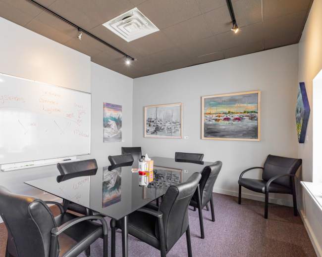 A conference room features a glass table surrounded by black chairs, with a whiteboard on one wall and two paintings hanging on another.
