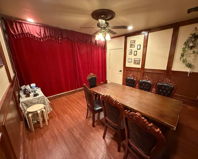 The image shows a dining room with a wooden table surrounded by eight ornate chairs, and a red curtain covering one wall.