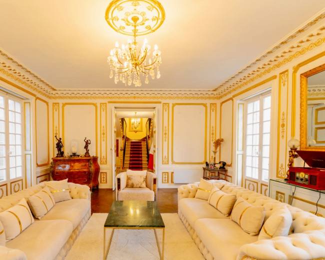 The room features two beige tufted sofas facing a coffee table, with ornate walls and a chandelier overhead, leading to a staircase at the back.