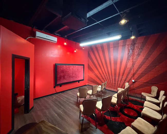 The image shows a small room with red walls featuring a graphic sunburst design, equipped with several chairs arranged in a semi-circle around a large screen.