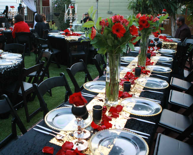 A beautifully arranged outdoor dining setup features black and gold tablecloths adorned with red flowers and rose petals, surrounded by elegantly set tables with black chairs.