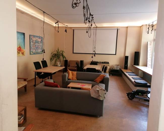 The image shows a spacious room with a gray sofa, a wooden table with chairs, and a projector screen, along with various audio equipment and decorative wall art.