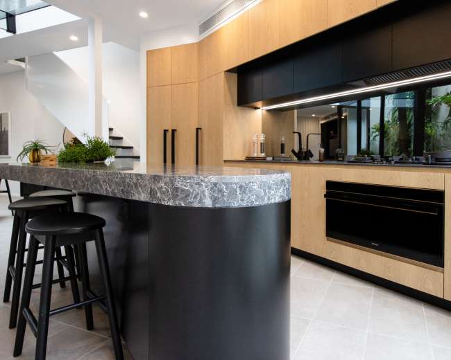 A modern kitchen features a marble countertop island with black stools, warm wood cabinetry, and sleek black appliances.