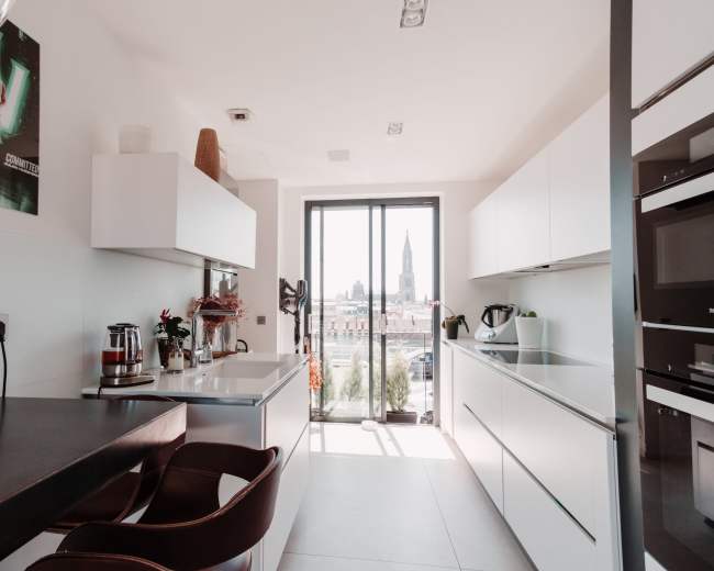 A modern kitchen features sleek white cabinetry, a small breakfast bar with two brown chairs, and a large window showcasing a cityscape with a prominent tower.