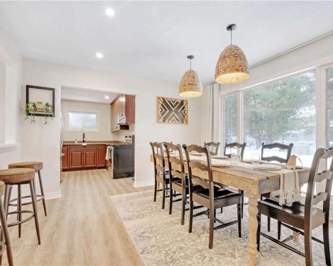 The image shows a bright dining area with a long wooden table surrounded by chairs, a kitchen visible in the background, and large windows letting in natural light.