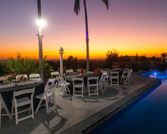 A beautiful outdoor setting features tables arranged near a pool as the sun sets, casting vibrant colors across the sky.